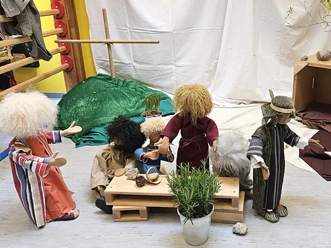 Puppen, die um einen Tisch versammelt sind. Im Hintergrund ein Kreuz.