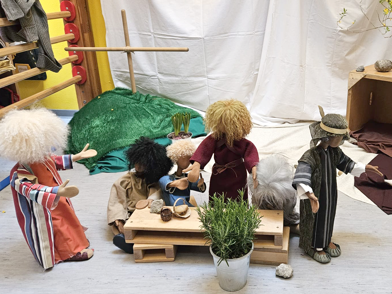 Puppen, die um einen Tisch versammelt sind. Im Hintergrund ein Kreuz.